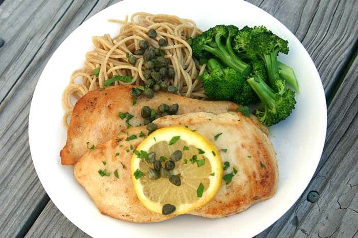 Chicken Scallopini on a white plate topped with a lemon slice and capers next to whole wheat spaghetti and steamed broccoli