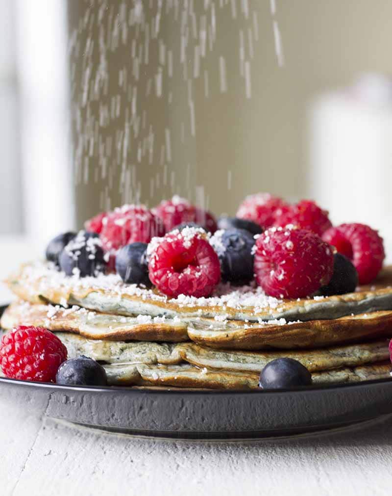 Sprinkling stevia over a stack of pancakes with berries and sugar free syrup on top