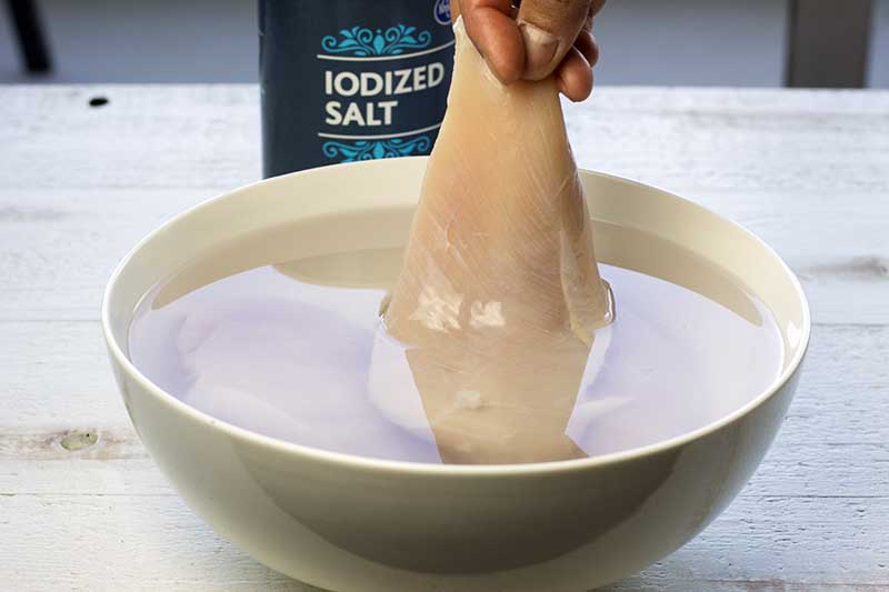 A chicken breast being lowered into a bowl with water and salt