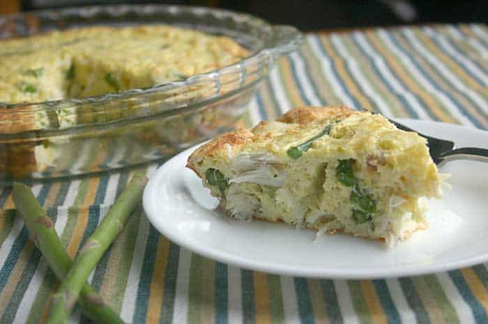 A slice of Crustless Crab and Asparagus Quiche on a white plate in front of a tray with the rest of the quiche