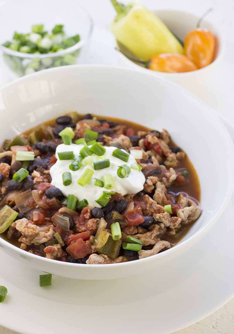 Turkey chili in a bowl, topped with sour cream and green onions. In the background, small bowls of chopped green onion and whole peppers