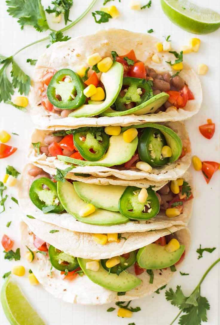 Four vegan pinto bean tacos on a plate with extra cilantro, tomatoes, corn, and lime wedges for decoration, as seen from above