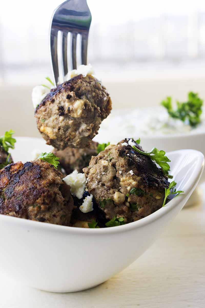 A bowl of bison meatballs topped with parsley, feta, and homemade tzatziki with one meatball on a fork
