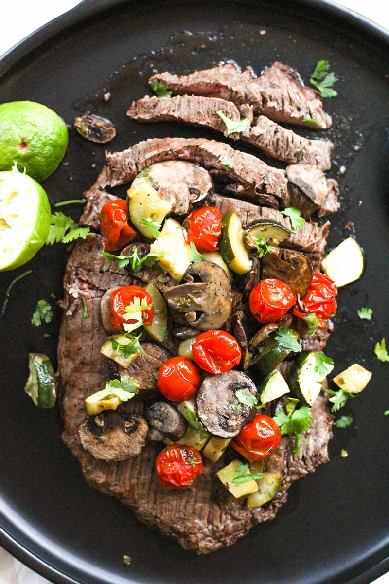 Marinated Flank Steak on a plate, garnished with zucchini, tomato, and mushroom with lime on the side