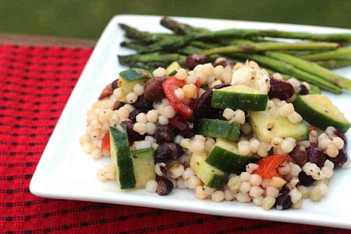 Pearl couscous salad with beans on a square white plate with asparagus over a red woven cloth placemat