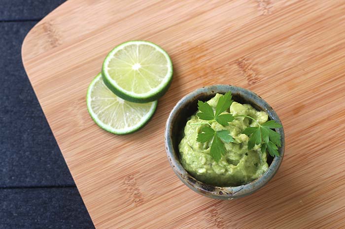 5-Minute Lime-Avocado Sauce topped with a sprig of parsley in a small serving ramekin on a wooden cutting board next to two lime slices