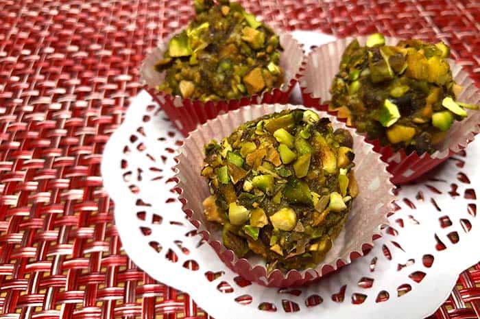 Three Key Lime Date Balls with Pistachios in truffle wrappers on top of a white doily on a red decorative woven placemat.