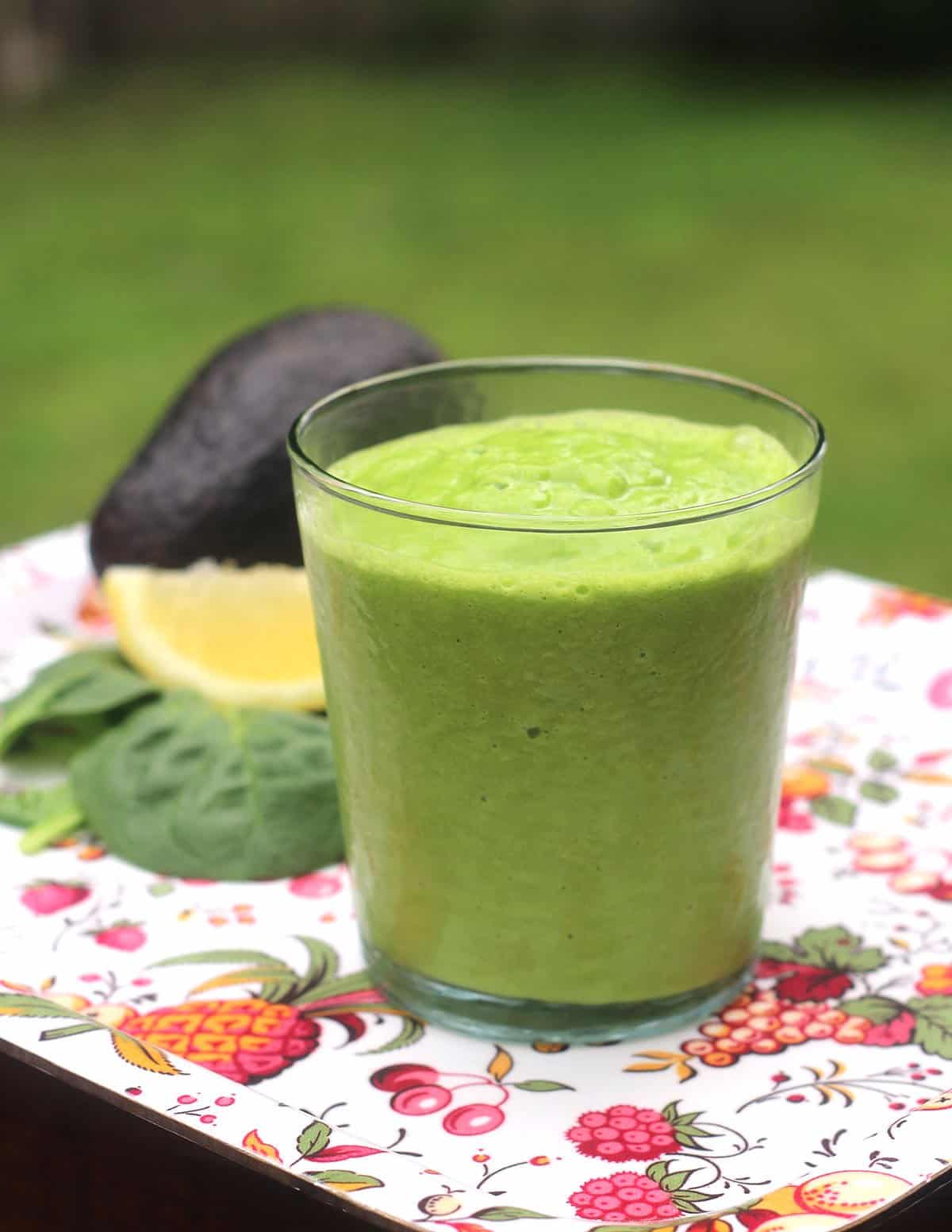 Avocado Smoothie with Leafy Greens in a glass with spinach, lemon, and an avocado in the background