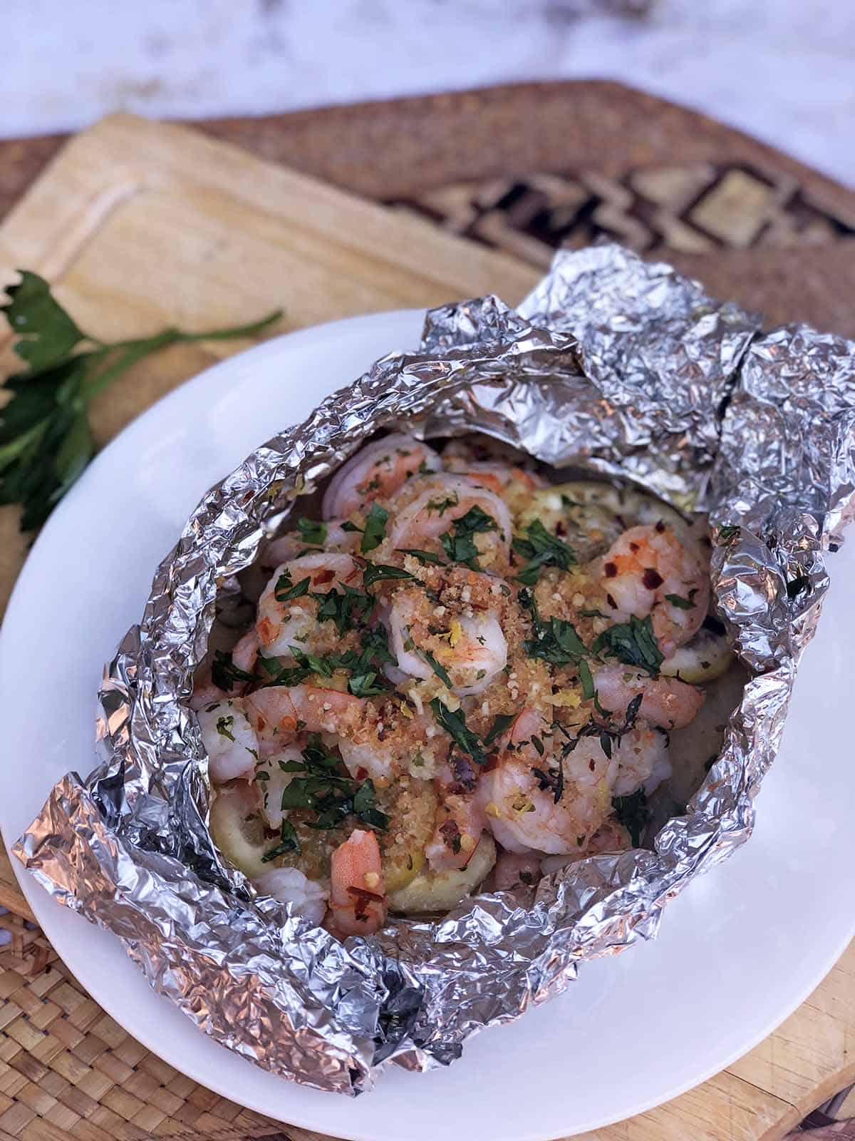 Cooked Shrimp Scampi in an open foil packet topped with bread crumbs and fresh parsley on a plate that's on top of a cutting board
