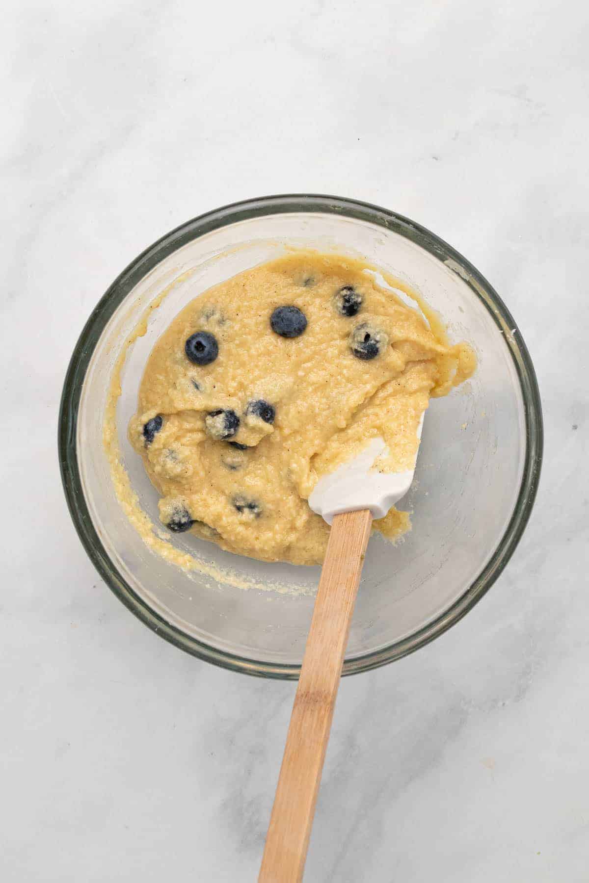 Muffin batter in a glass mixing bowl with blueberries folded in and a rubber spatula