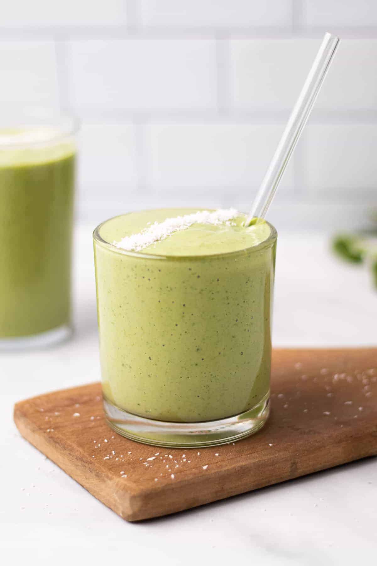 Green smoothie in a glass with a straw, topped with fine coconut, sitting on a wooden cutting board
