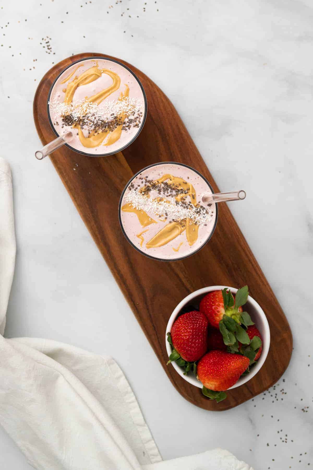 Two smoothies on a wooden board with a bowl of strawberries