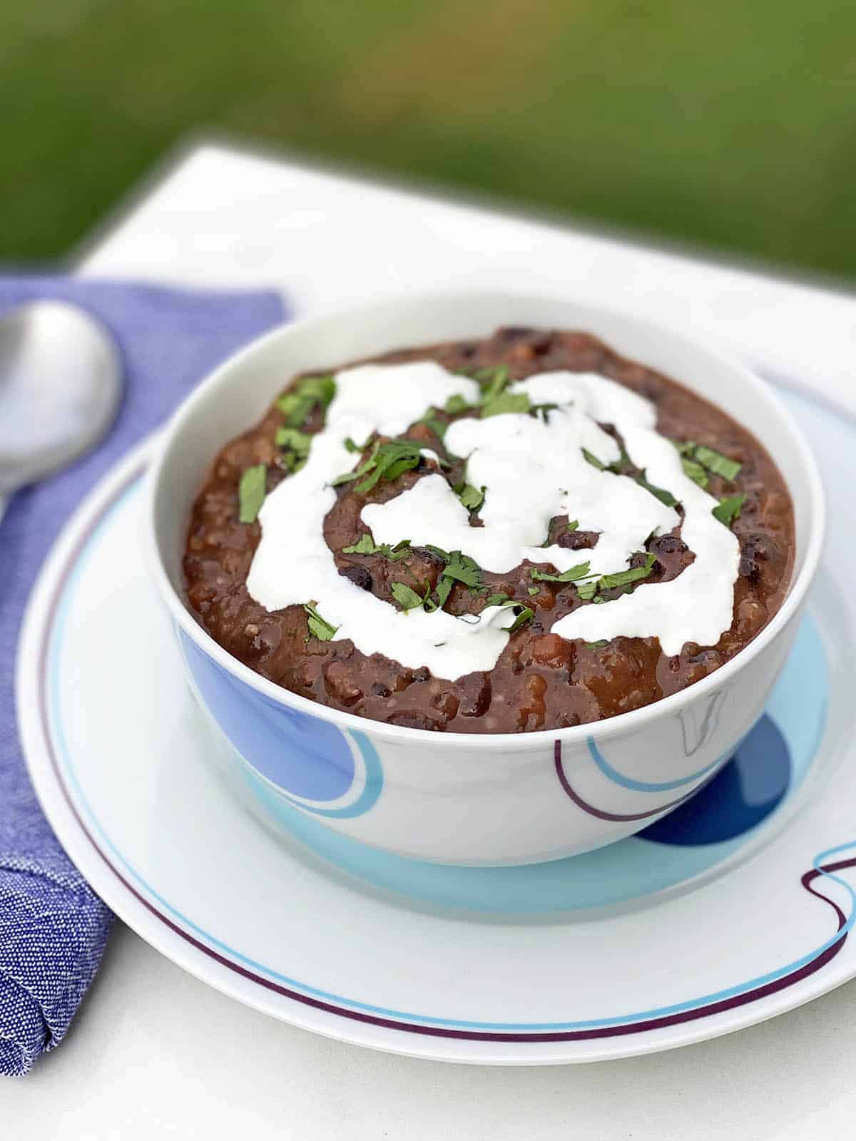 Black Bean Soup with Cilantro-Lime Cream in a white bowl on a white plate with a blue napkin and spoon next to it
