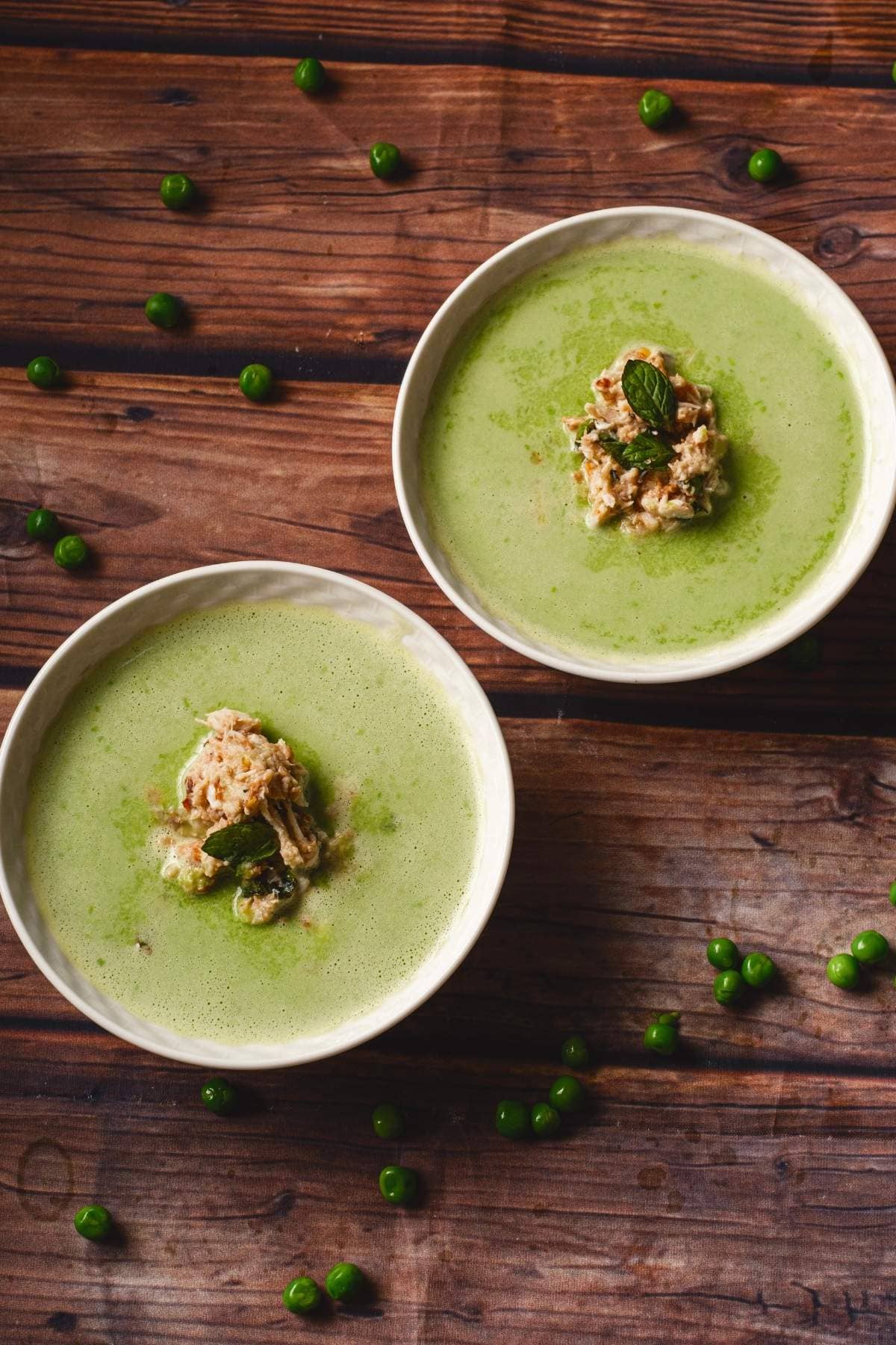 Two cups of soup surrounded by peas on a wooden table 