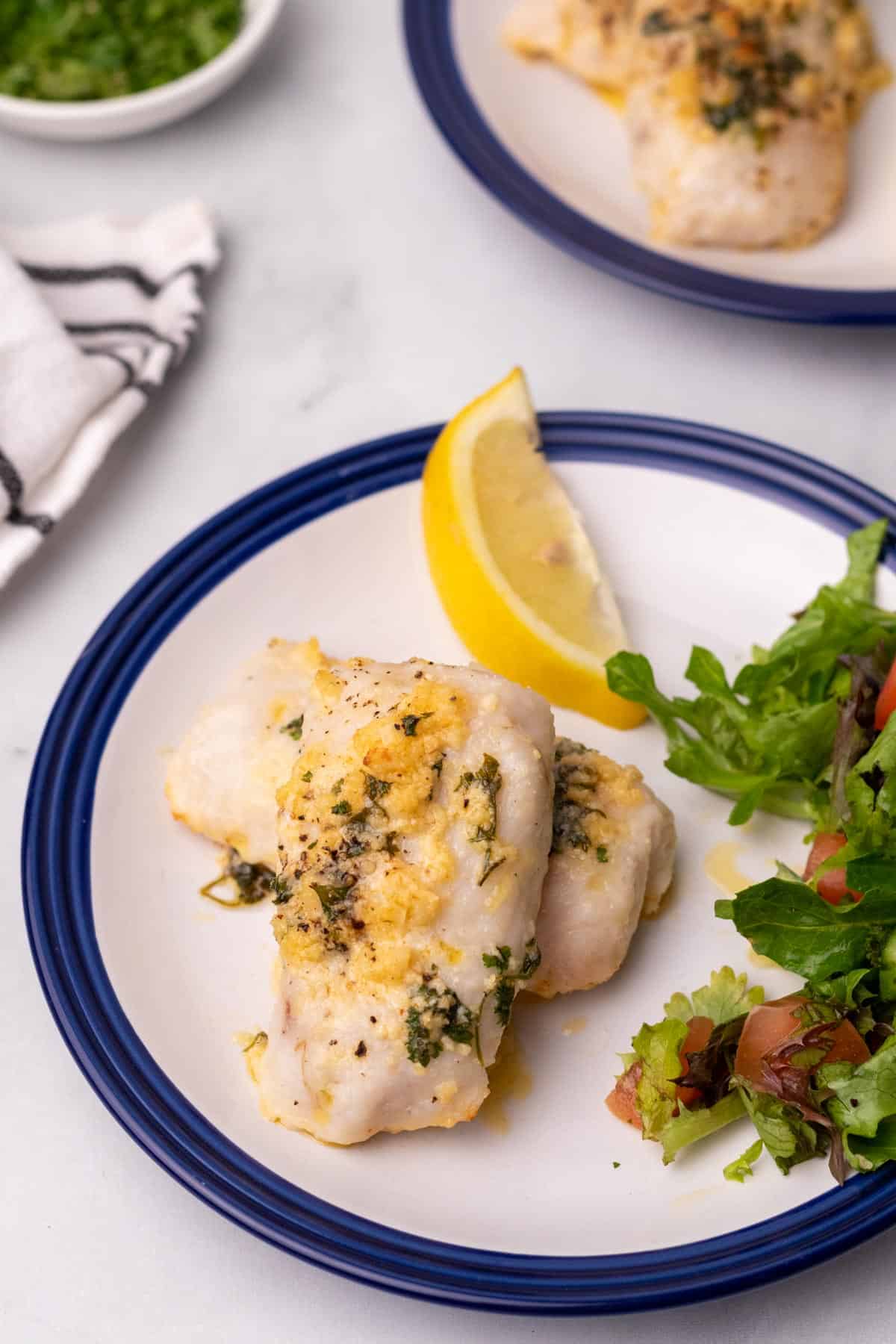 Two cod filets on a plate with a lemon wedge and a salad