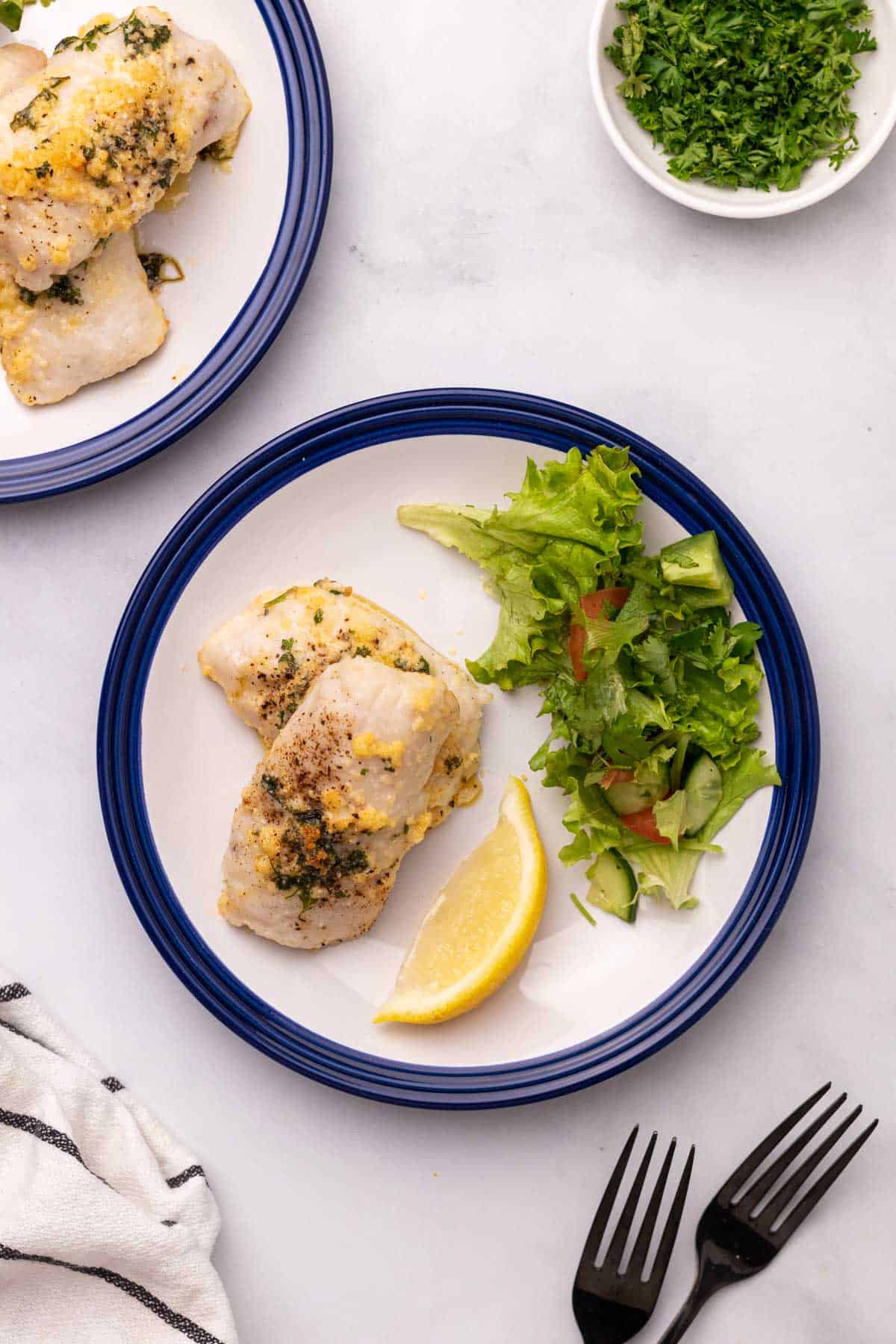Two cod filets on a plate with a lemon wedge and salad