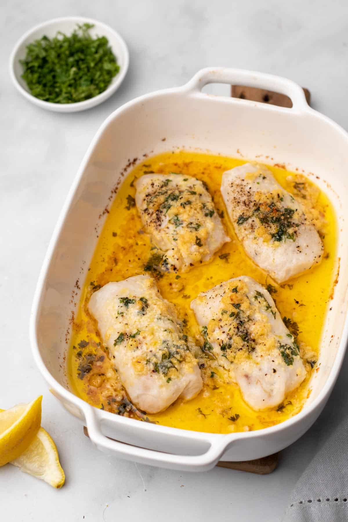 Baked cod with parmesan and garlic butter in a baking dish next to a ramekin of parsley and lemon wedges