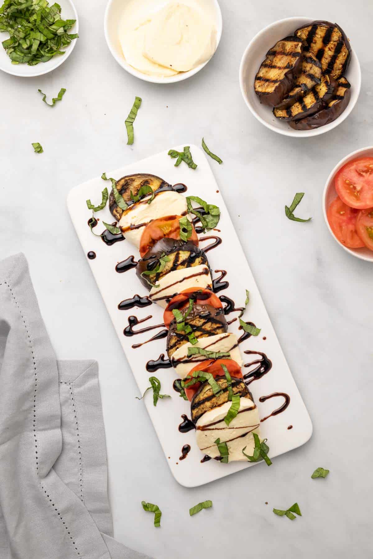 Caprese salad with eggplant on a white serving dish, as seen from above, next to bowls with individual ingredients