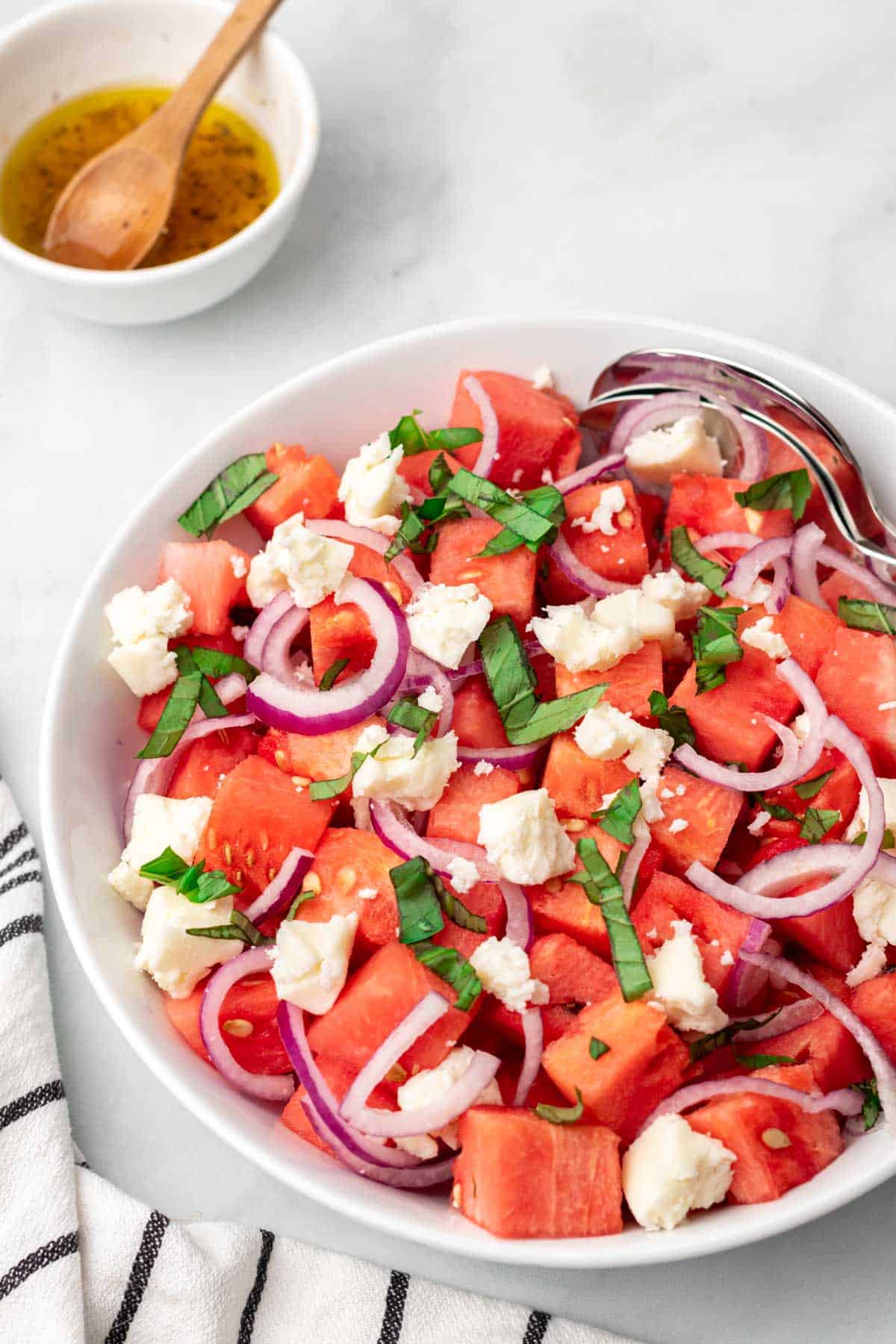 Salad with watermelon, onion, goat cheese, and basil in a white bowl next to a ramekin with salad dressing