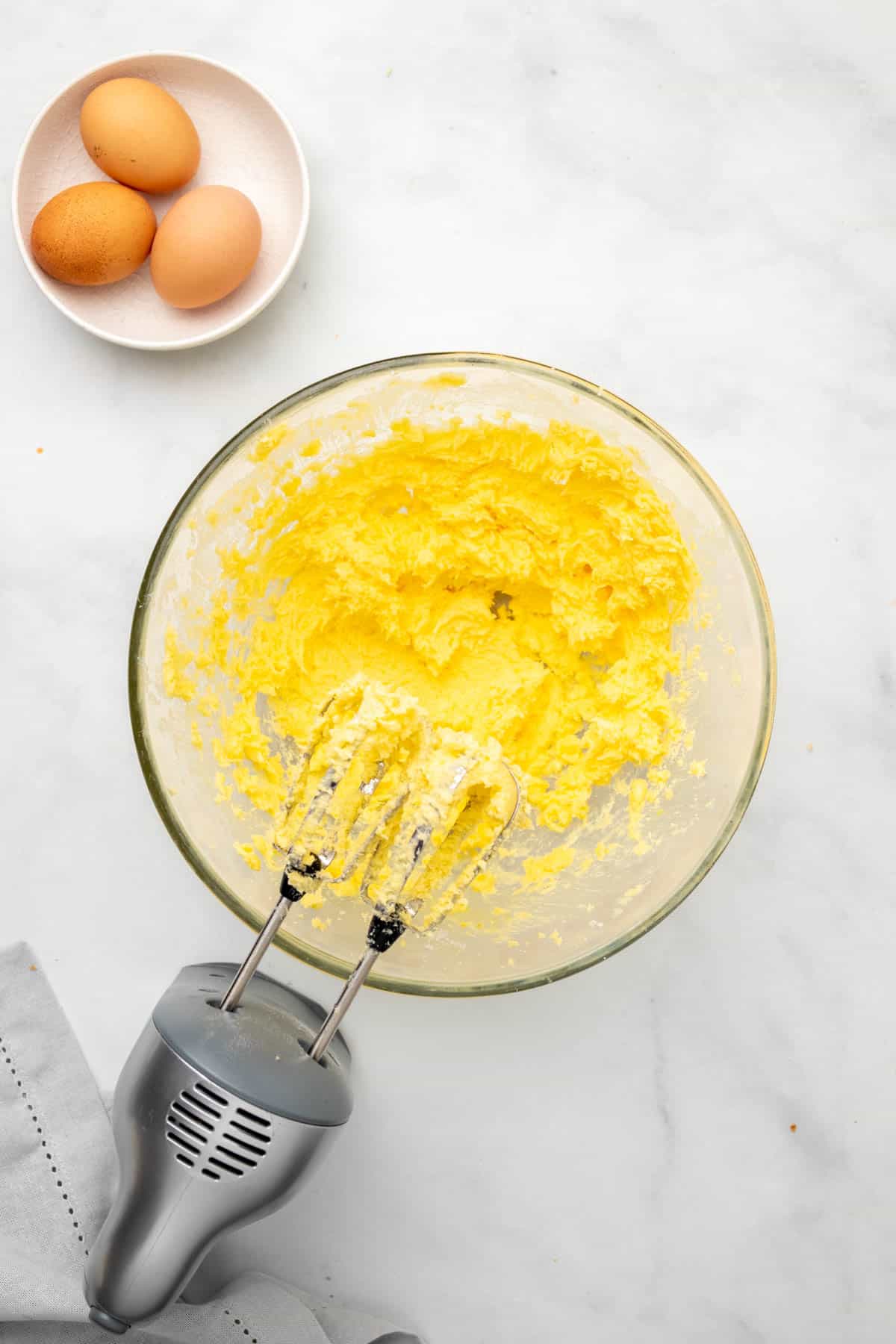 Pound cake batter mixed with hand beaters in a glass bowl next to a ramekin with 3 eggs