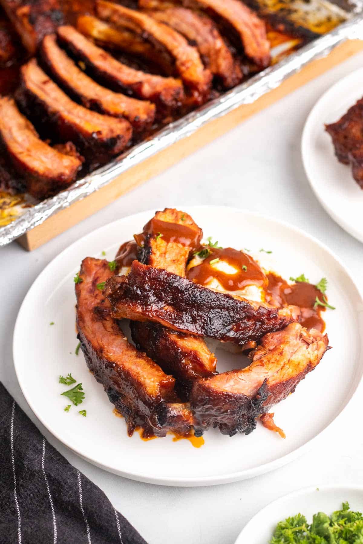 Four ribs on a white plate next to a tray of sliced ribs