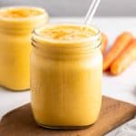 Carrot smoothie in a small glass mug with a glass straw on top of a wooden serving tray