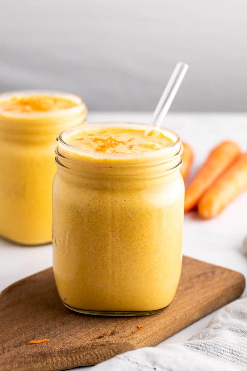 Carrot smoothie in a small glass mug with a glass straw on top of a wooden serving tray