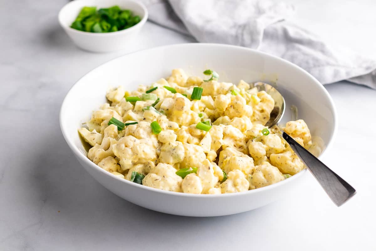 Cauliflower potato salad in a white bowl with a spoon, garnished with fresh green onion