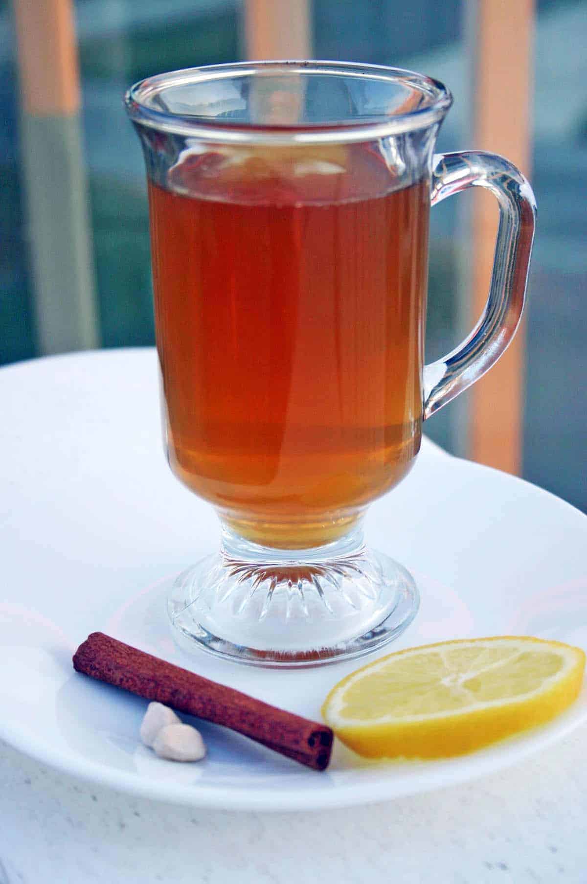 Spiced Ginger Tea in a glass mug on a white plate next to a slice of lemon, a cinnamon stick, and cardamom pods