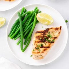 Yogurt marinated chicken breast on a white plate with steamed green beans and a wedge of lemon