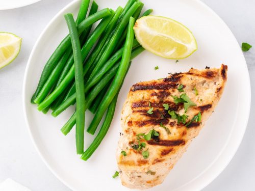 Yogurt marinated chicken breast on a white plate with steamed green beans and a wedge of lemon
