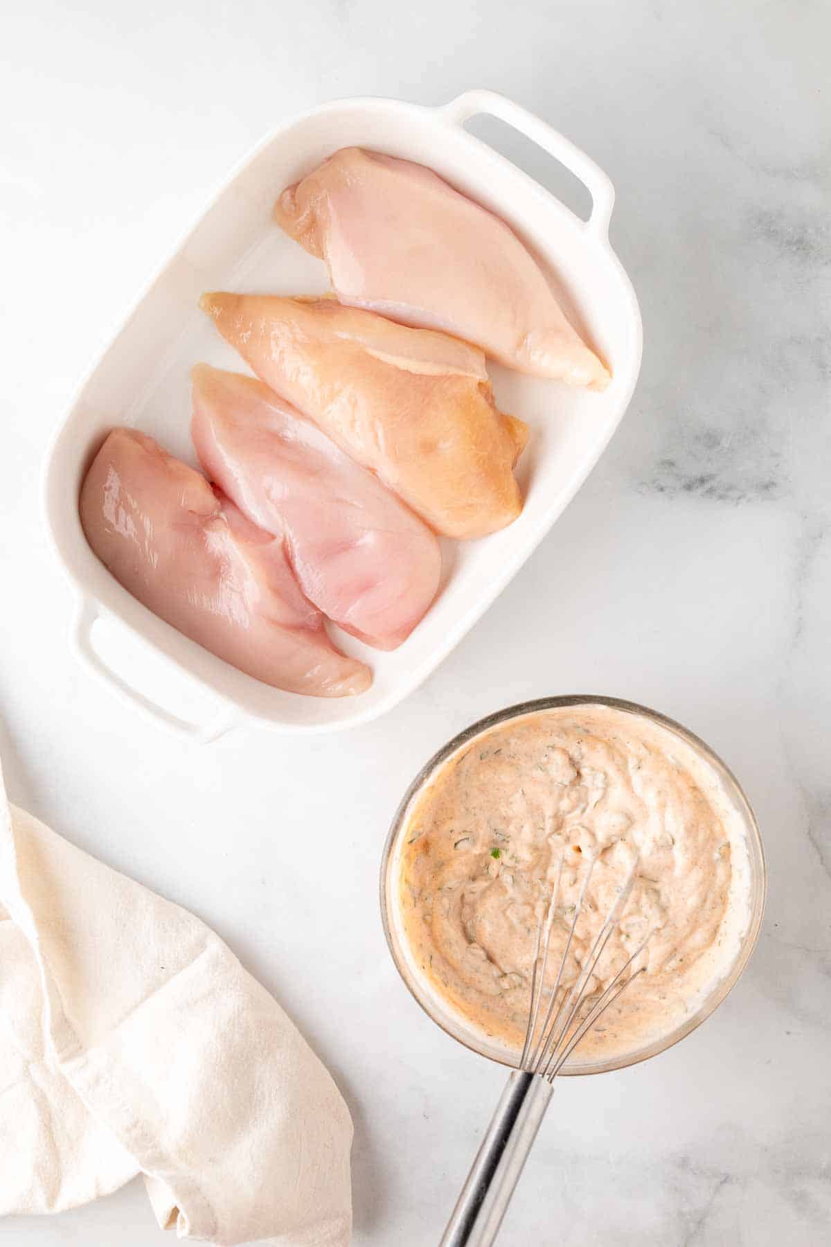 Four chicken breasts in a baking dish next to a glass dish with the marinade