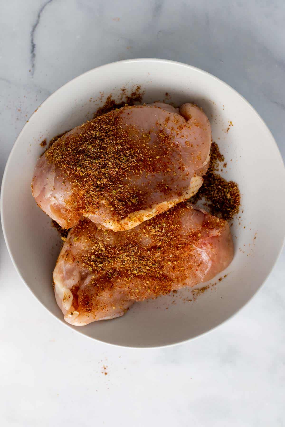 Chicken breasts in a white bowl, sprinkled with seasoning mixture