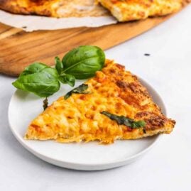 A slice of keto chicken crust pizza on a white plate next to basil leaves
