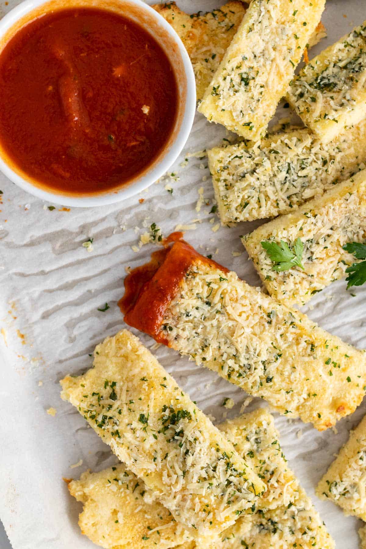 Strips of keto garlic bread on a serving tray with marinara sauce; one slice has been dipped in the marinara; as seen from above