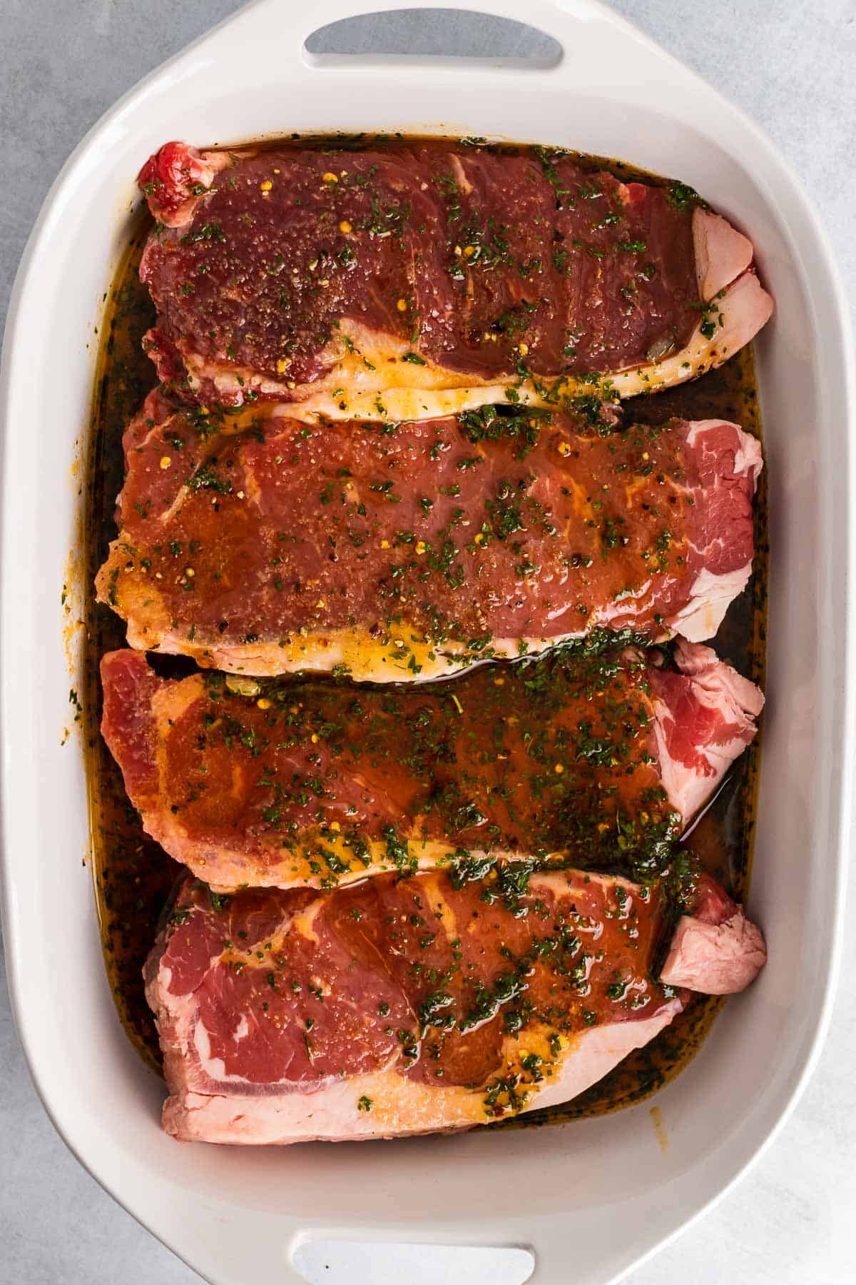 Four steaks marinating in a large white dish