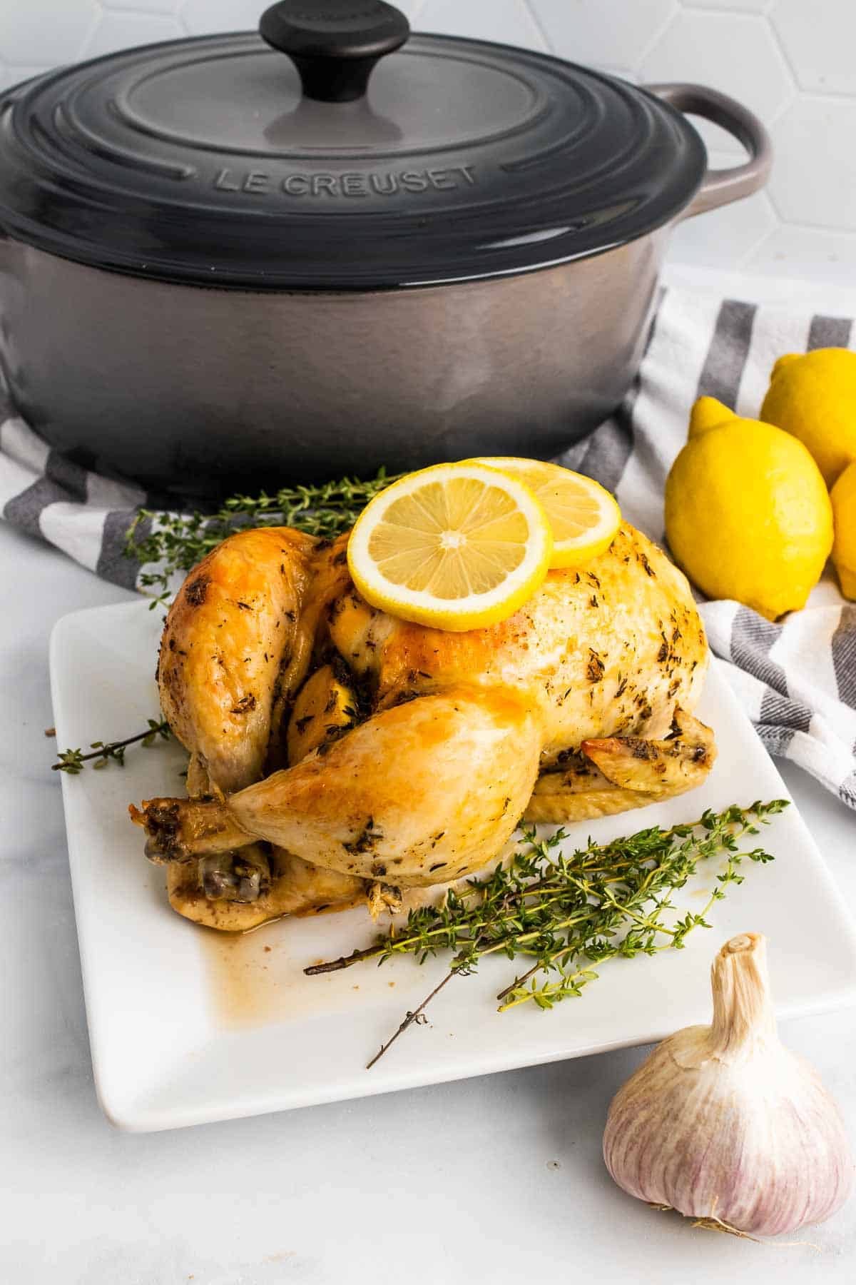 Dutch oven chicken on a white plate next to thyme sprigs and garnished with two lemon slices