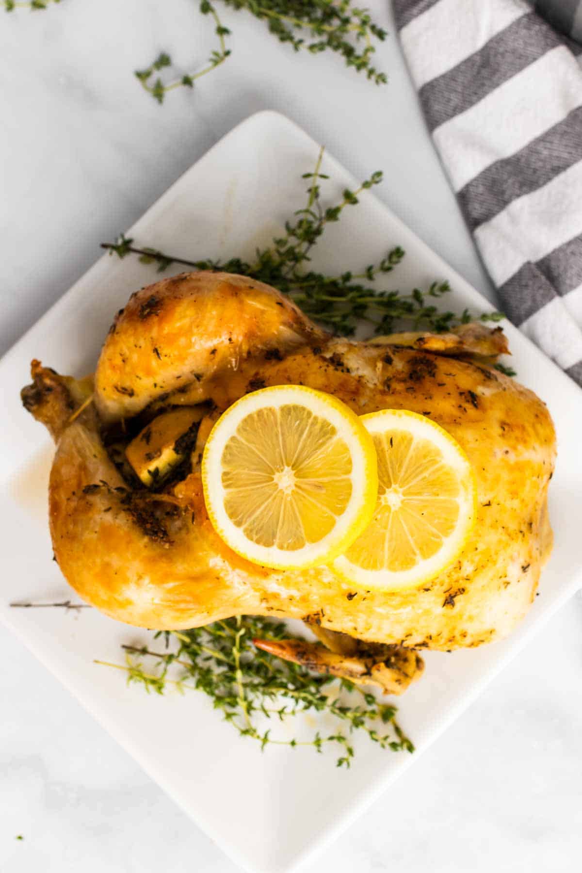 Whole chicken on a white plate next to sprigs of thyme and garnished with two lemon slices, as seen from above