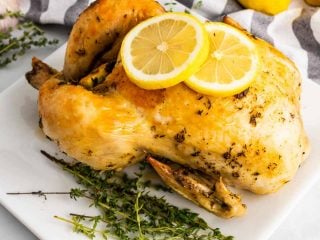 Dutch oven chicken on a white plate next to thyme sprigs and garnished with two lemon slices