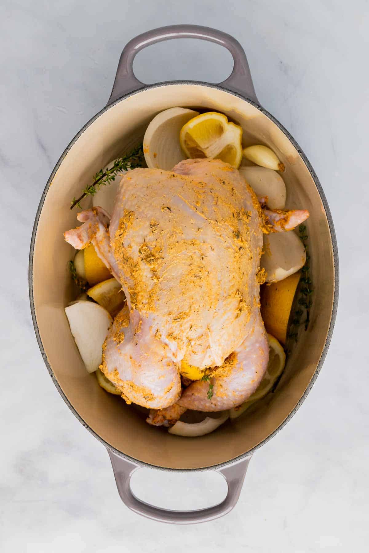 Buttered chicken placed on top of onions, lemons, and herbs in a dutch oven, as seen from above