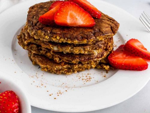 A stack of four flourless banana pancakes on a white plate, topped with and surrounded by strawberry slices