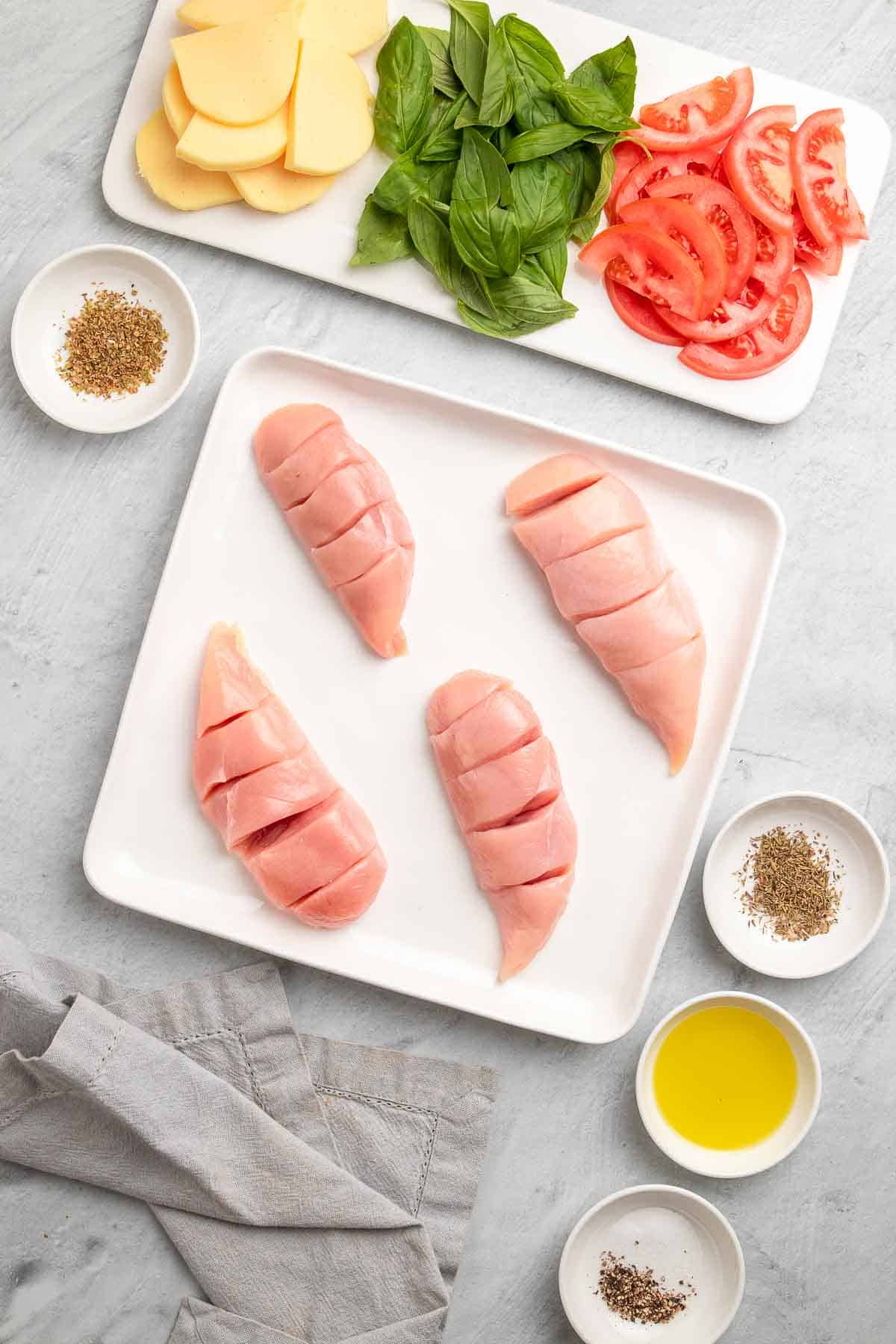 Four sliced chicken breasts on a white plate next to a white serving tray and four ramekins with the rest of the ingredients