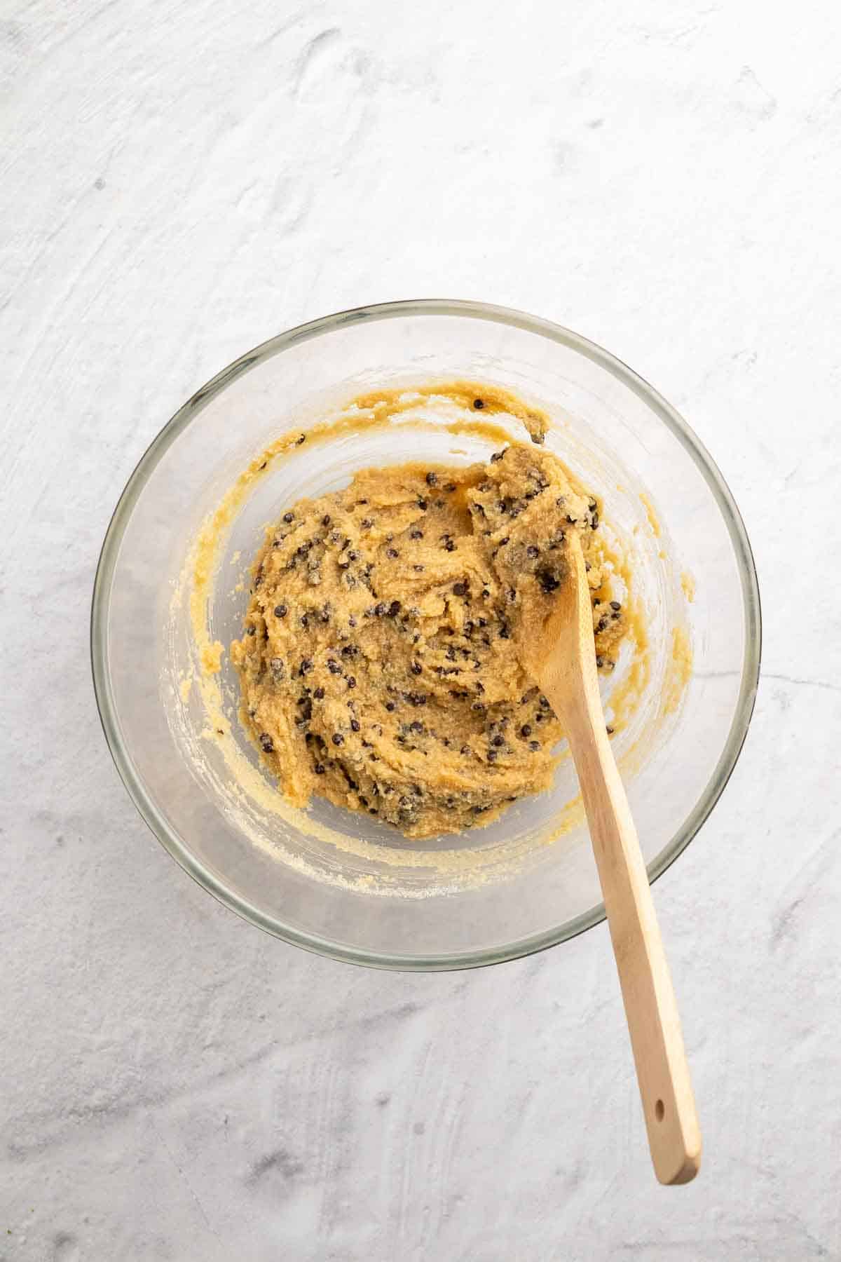 Chocolate chips folded in to the cookie dough in a glass bowl with a wooden spoon, as seen from above