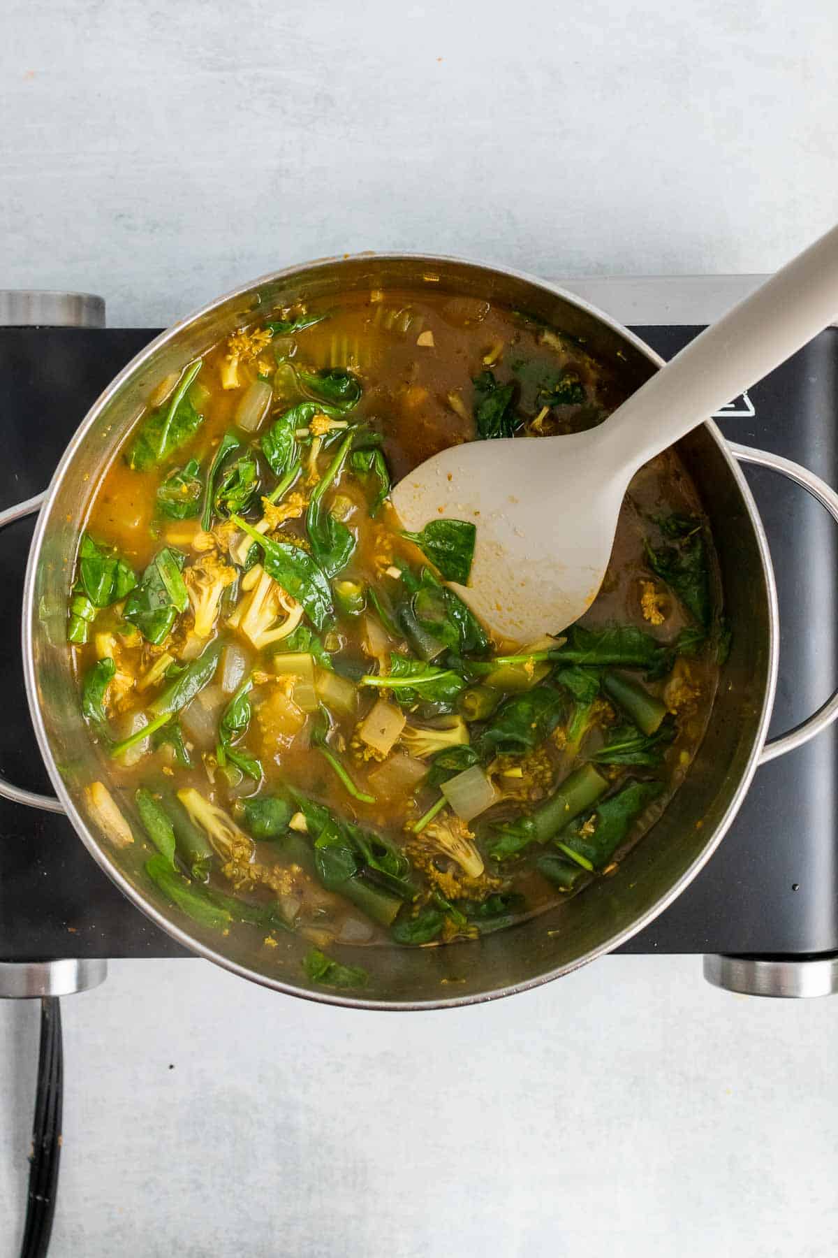 A white spoon stirring the spinach into the soup in the saucepan, as seen from above