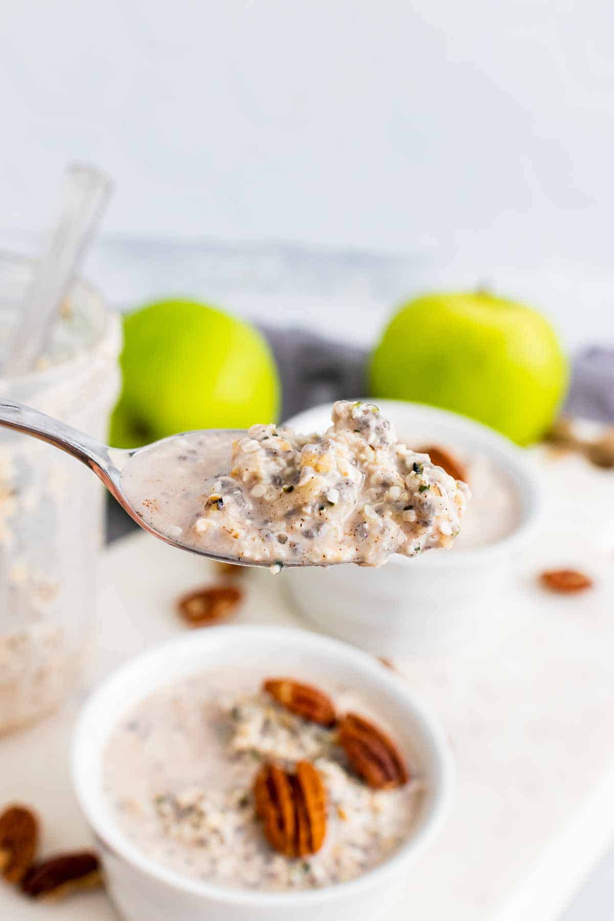 A spoonful of oats with two bowls of oats topped with pecans, an empty glass jar, and two apples in the background
