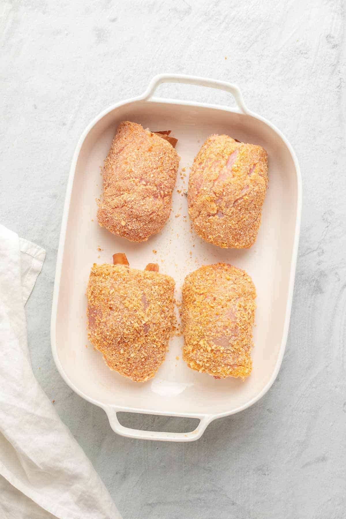 Four breaded and rolled chicken breasts in a white baking dish, as seen from above