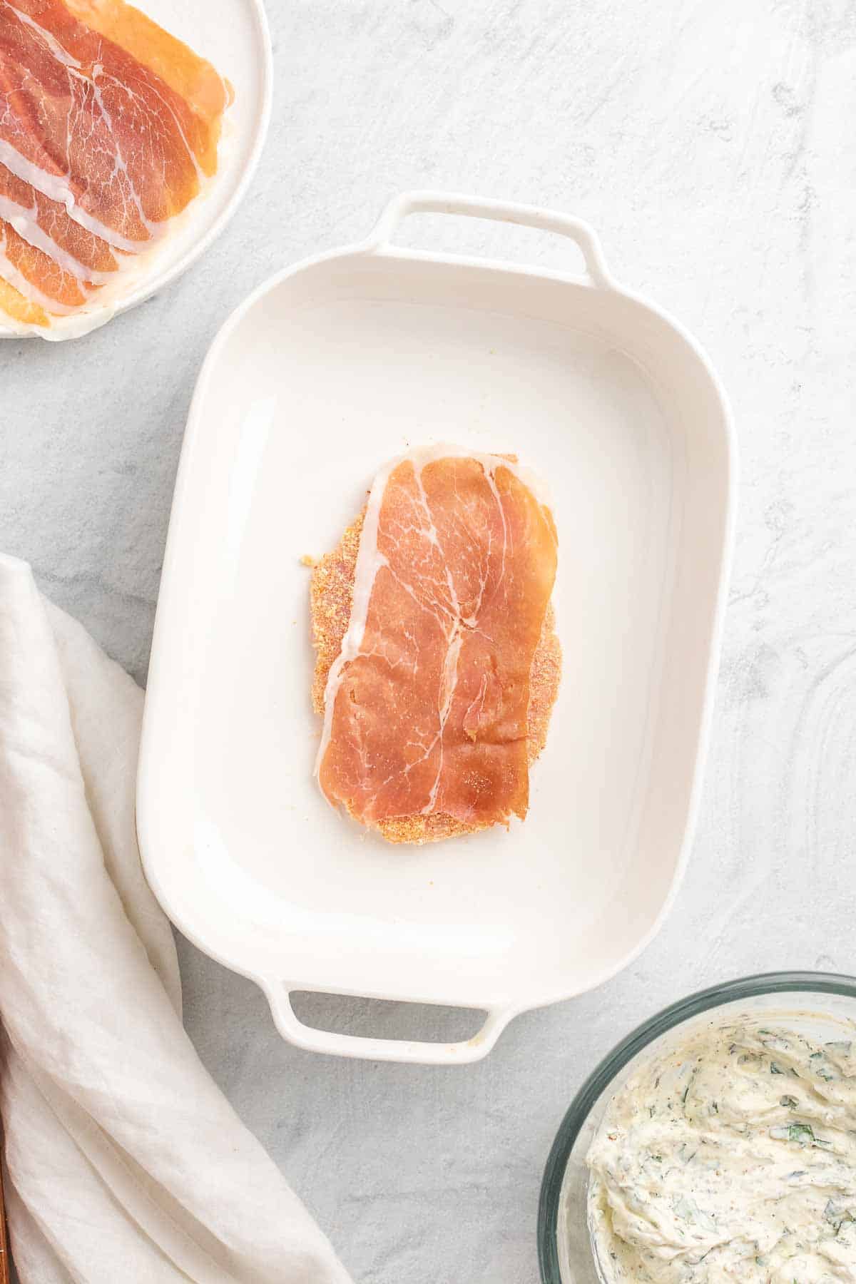 Flattened and coated chicken breast set in a baking dish with a slice of prosciutto spread out over the breast, as seen from above