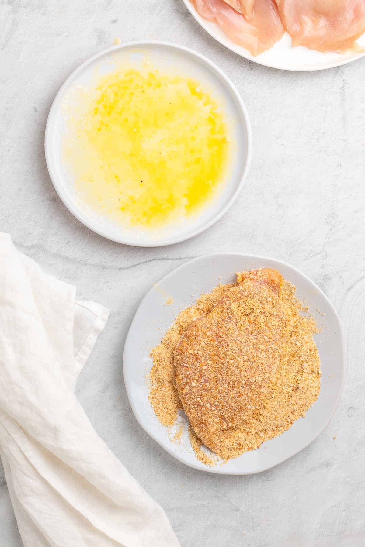 Flattened chicken breast on a white plate of low-carb breadcrumbs next to a white plate of the lemon and butter mixture, as seen from above