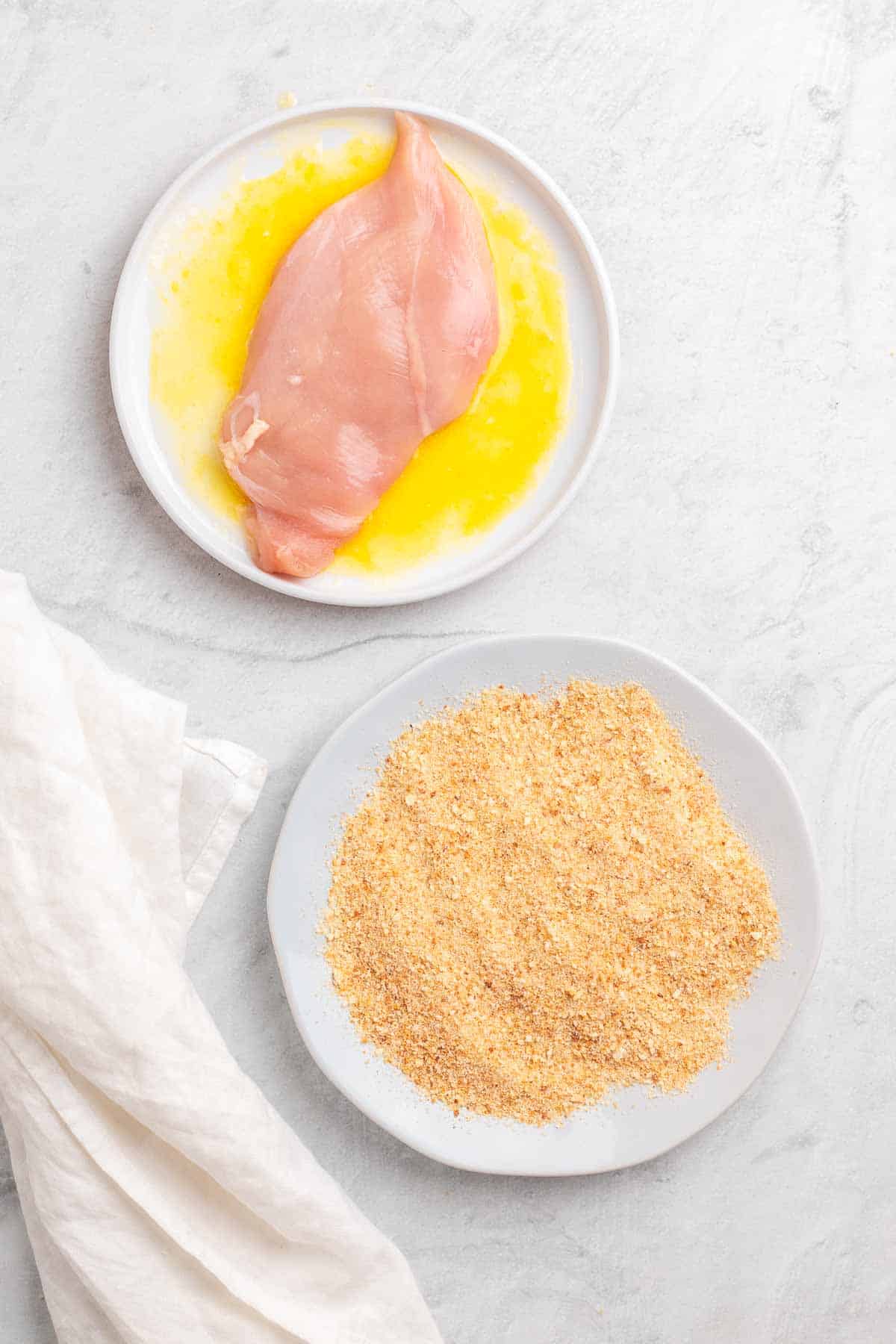 A flattened chicken breast in the lemon and butter mixture on a white plate next to another white plate of low-carb breadcrumbs, as seen from above