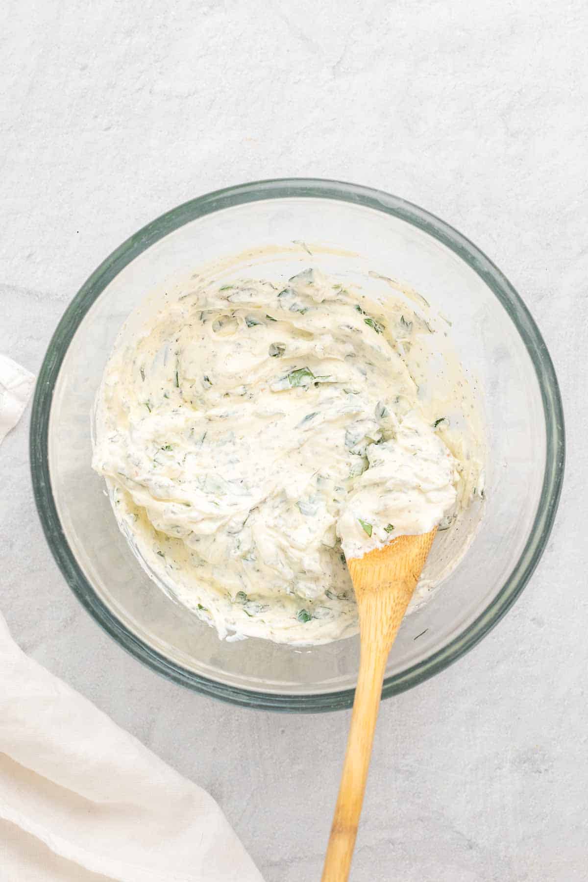 Ricotta, herbs, and seasoning mixed together in a large glass bowl with a wooden spoon, as seen from above